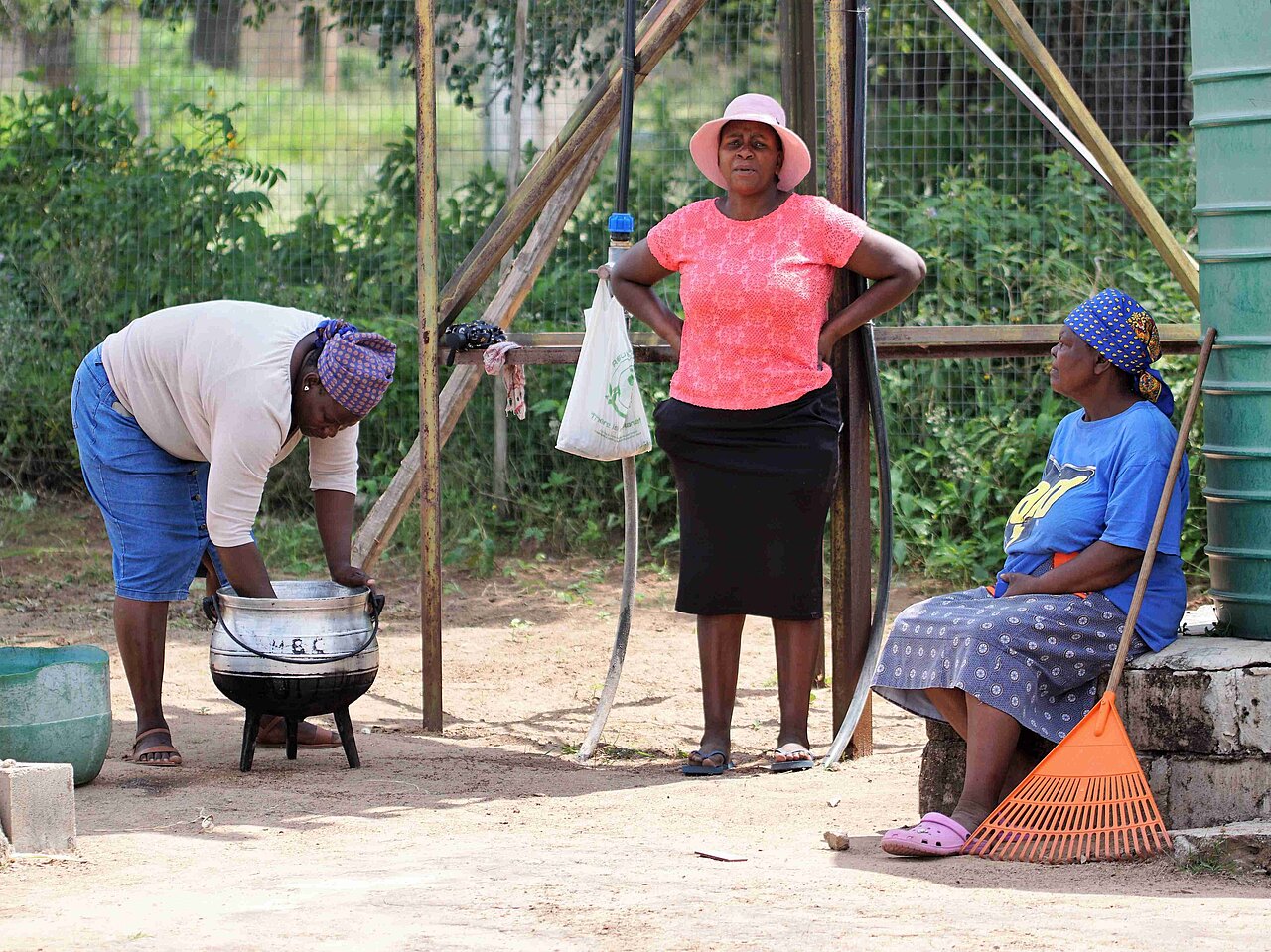 Drei Frauen der Gemeinde Welverdiend in Südafrika sitzen im Schatten, eine Frau reinigt einen Kessel