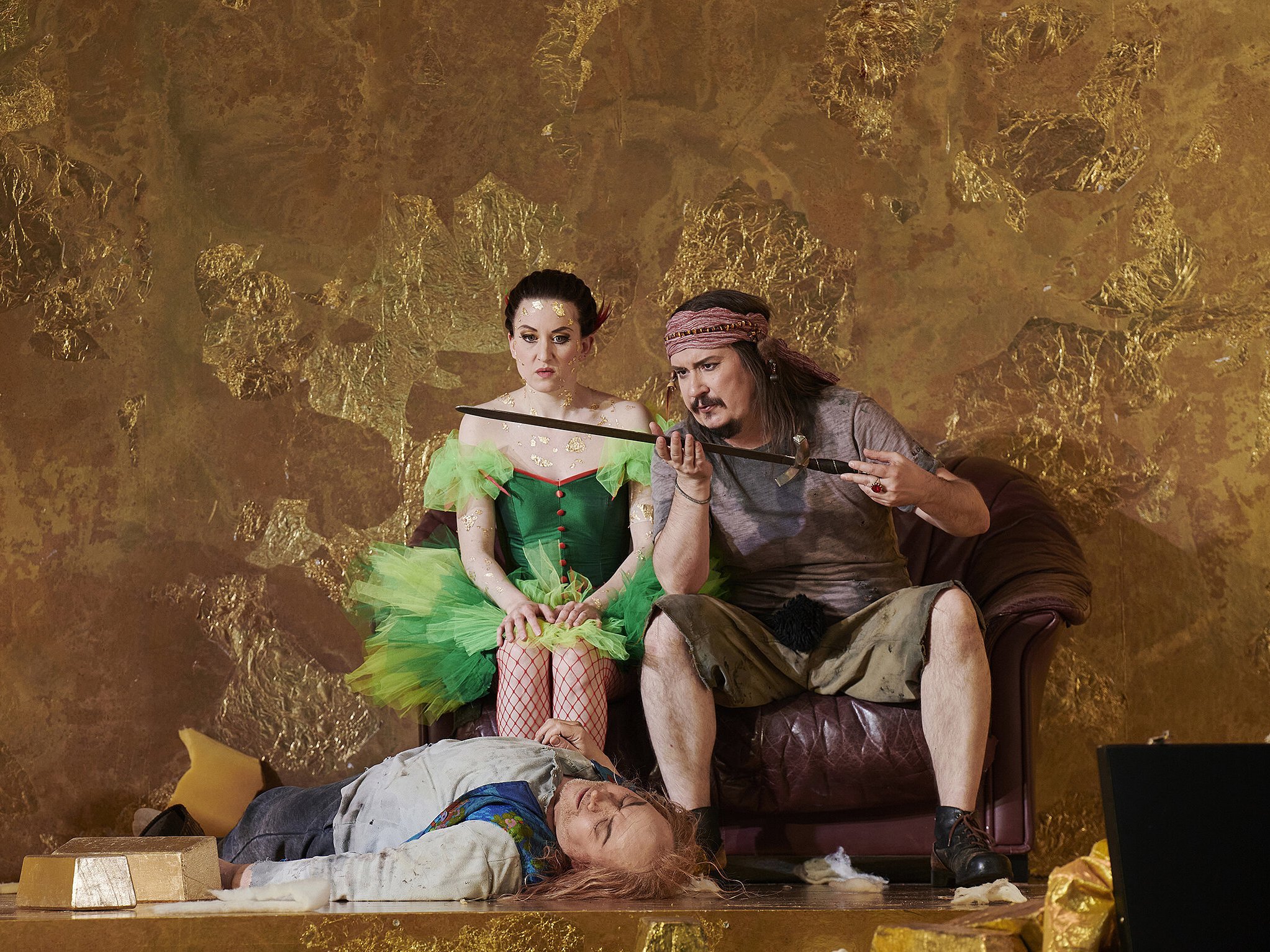 Zwei Schauspieler (links im grünen Feenkleid, rechts in T-Shirt und Shorts mit einem Schwert in der Hand) sitzen vor einer goldenen Wand, während eine Person vor ihnen auf dem Boden liegt