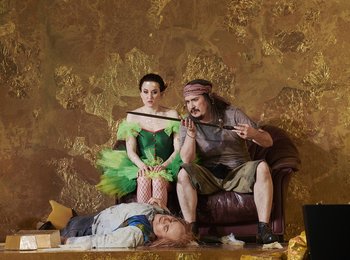 Zwei Schauspieler (links im grünen Feenkleid, rechts in T-Shirt und Shorts mit einem Schwert in der Hand) sitzen vor einer goldenen Wand, während eine Person vor ihnen auf dem Boden liegt