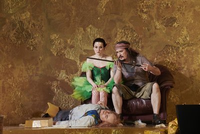 Zwei Schauspieler (links im grünen Feenkleid, rechts in T-Shirt und Shorts mit einem Schwert in der Hand) sitzen vor einer goldenen Wand, während eine Person vor ihnen auf dem Boden liegt