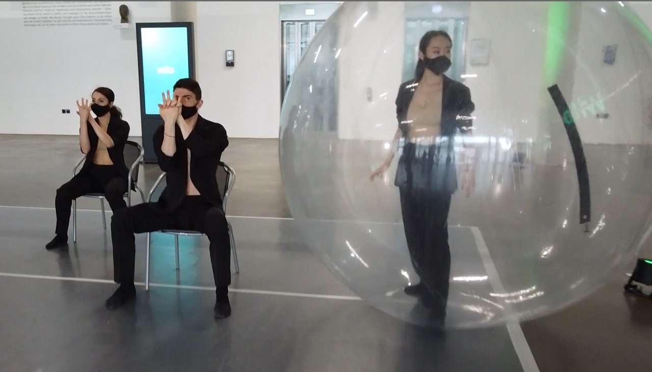 Künstlerinnen und Künstler des NRW Junior Balletts am Theater Dortmund, zwei Personen auf einem Stuhl, eine Person in einem transparenten Ballon