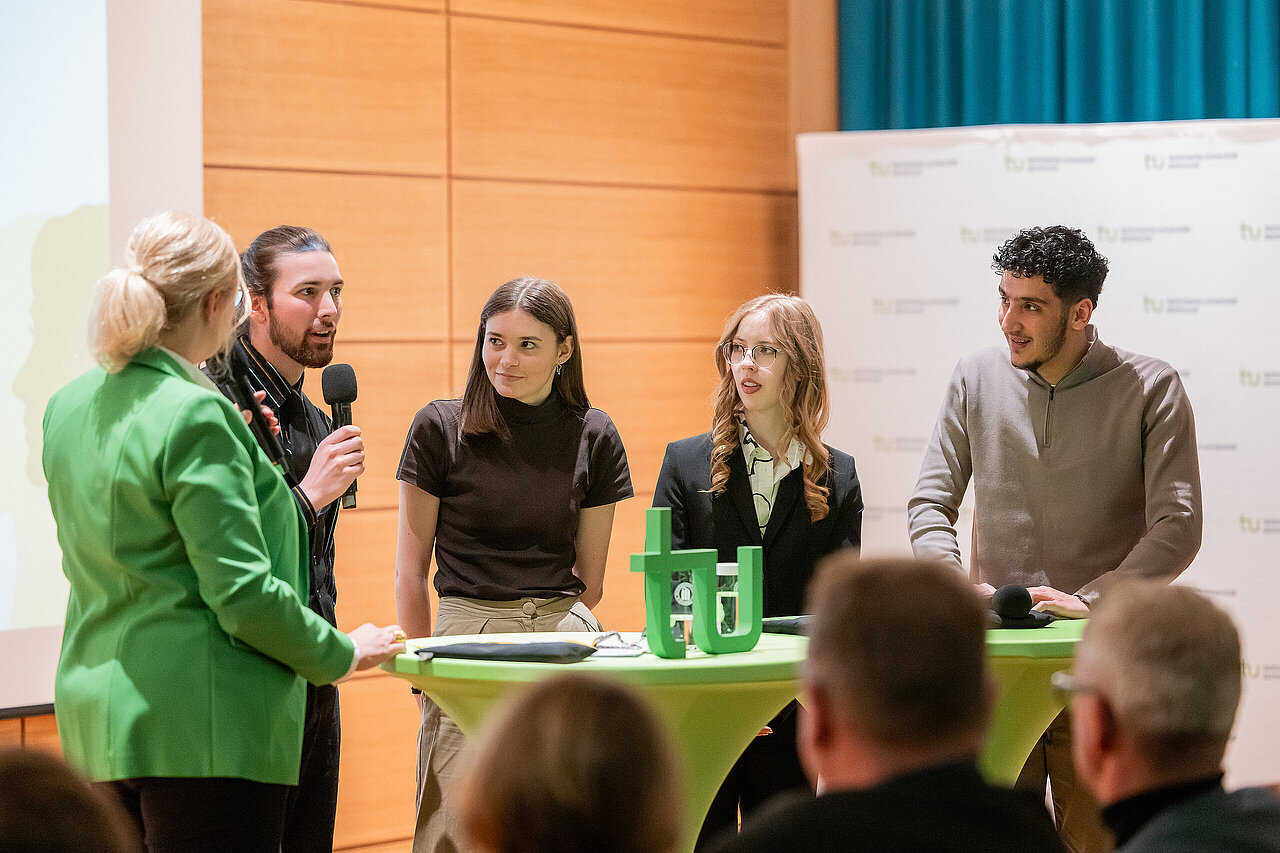 Vier Studierende stehen an einem Hochtisch und sprechen in ein Mikrofon