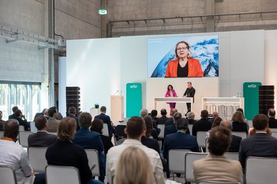 Oliver Hermes auf der Bühne, auf der Leinwand im Hintergrund spricht Svenja Schulze