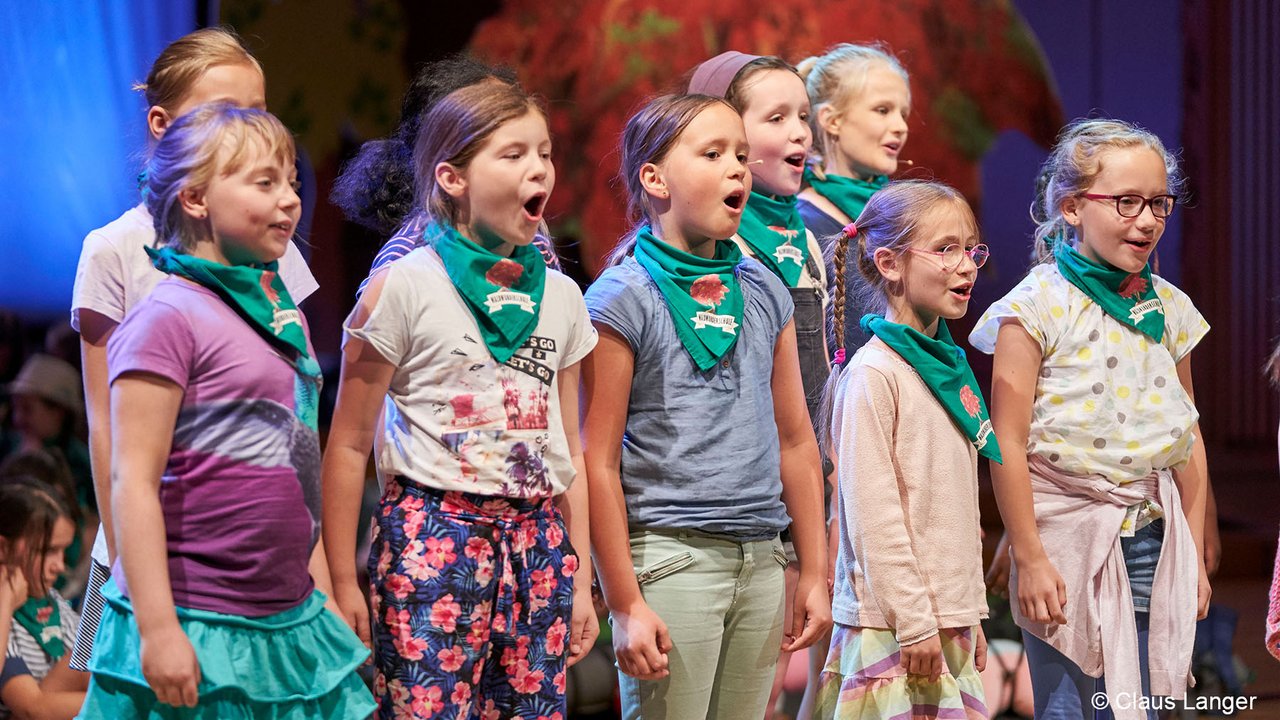 Mehrere Kinder mit grünen Halsbändern des WDR-Kinderchors stehen auf einer Bühne und singen