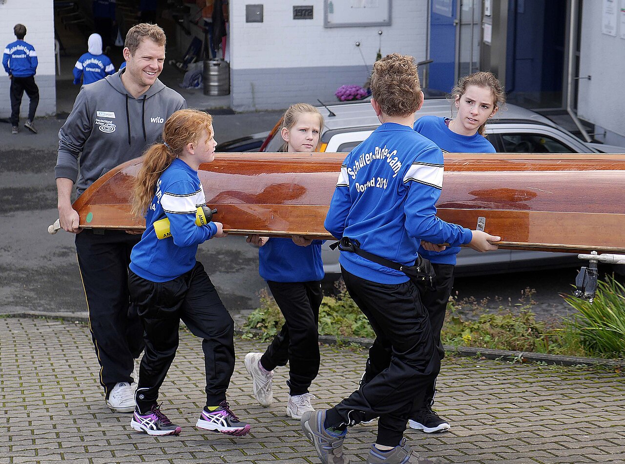 Kinder tragen mit Unterstützung des Team Deutschland-Achter ein Ruderboot zum Wasser