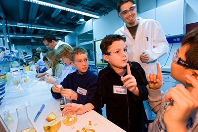 Drei Kinder mit Schutzbrillen an einem Experimentiertisch, zwei Kinder melden sich mit erhobenem Zeigefinger, hinter ihnen steht ein Doktorand in Forschungskittel