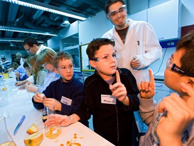 Drei Kinder mit Schutzbrillen an einem Experimentiertisch, zwei Kinder melden sich mit erhobenem Zeigefinger, hinter ihnen steht ein Doktorand in Forschungskittel