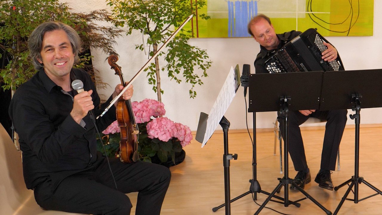 Zwei Musiker des Instituts für musikalische Bildung, im Vordergrund ein Mann mit Geige und Mikrofon, im Hintergrund ein Mann mit Akkordeon